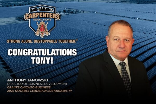 A man in a suit stands in front of a field of solar panels, with congratulatory text reading 