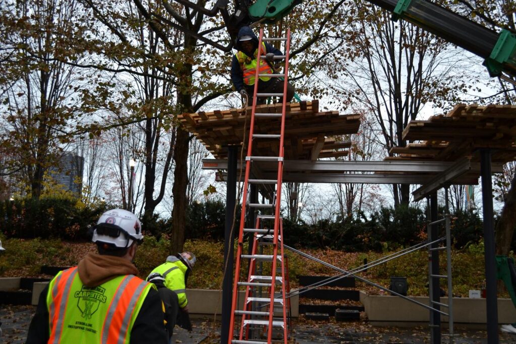 Several construction workers in safety vests and helmets work outdoors, with one standing on a ladder supporting wooden pallets on a metal frame. Trees with autumn leaves are in the background.